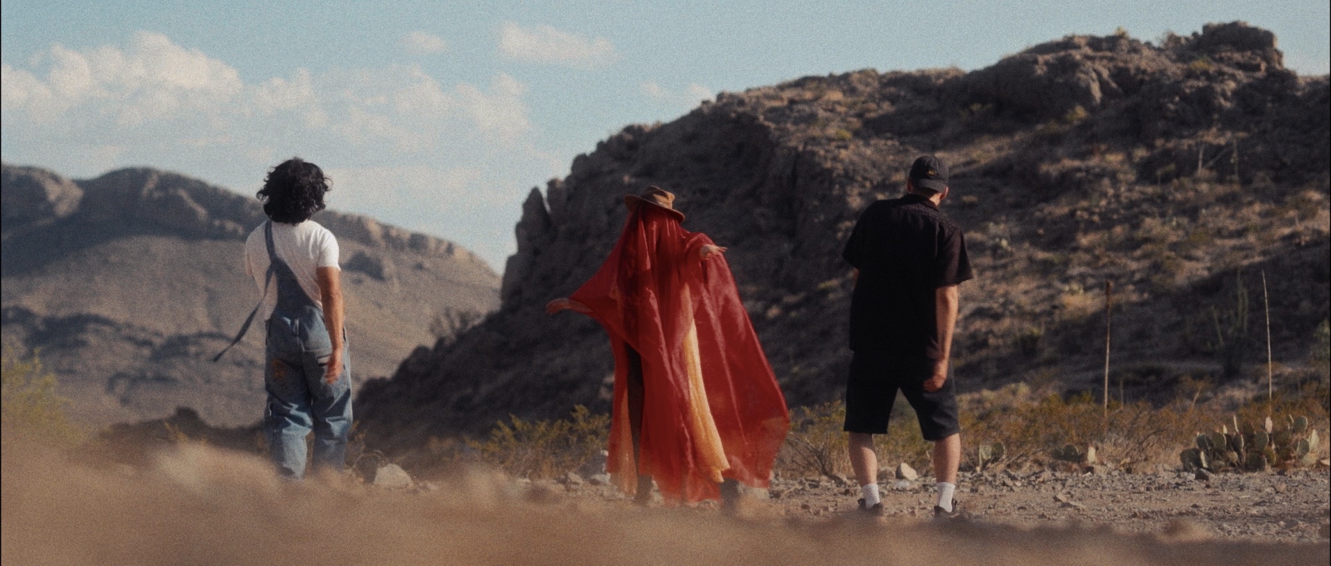 Fotograma de Querida Flaca: una figura cubierta de rojo aparece entre dos hombres en el desierto